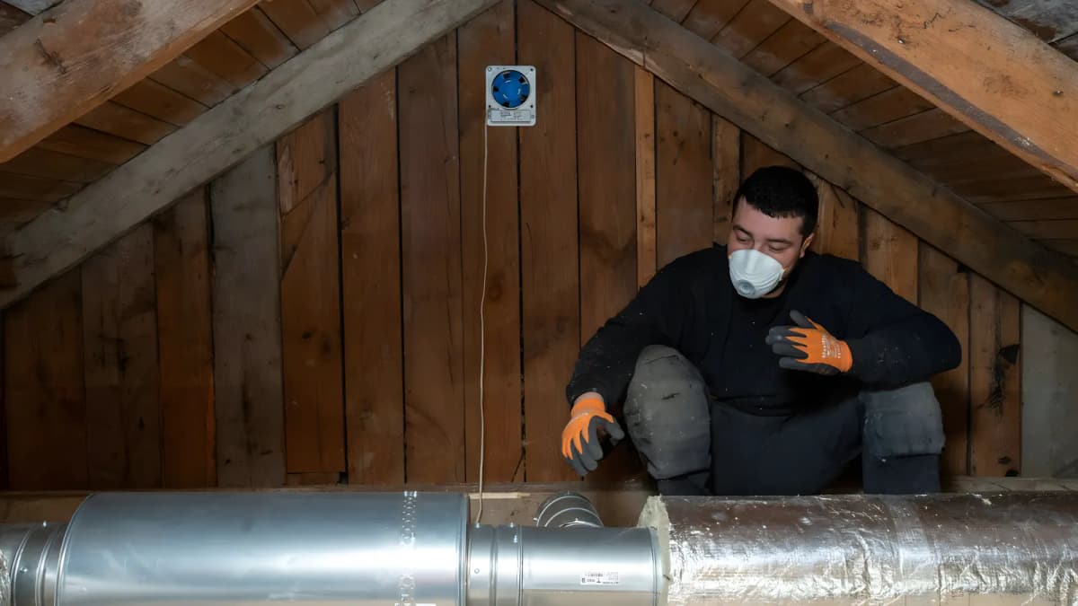 Montör med andningsskydd och handskar sitter på en vind i Stockholm bakom blanka ventilationskanaler