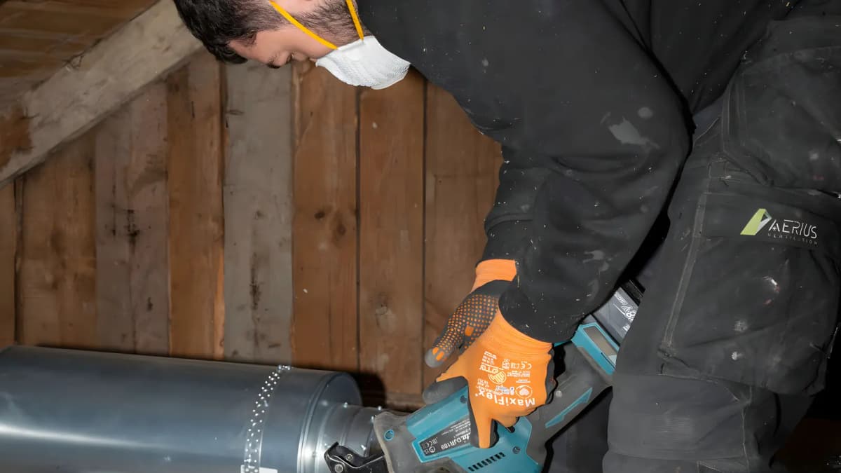 Tekniker med andningsskydd och orangea handskar skruvar ihop en galvaniserad ventilationskanal på en vind i Stockholm